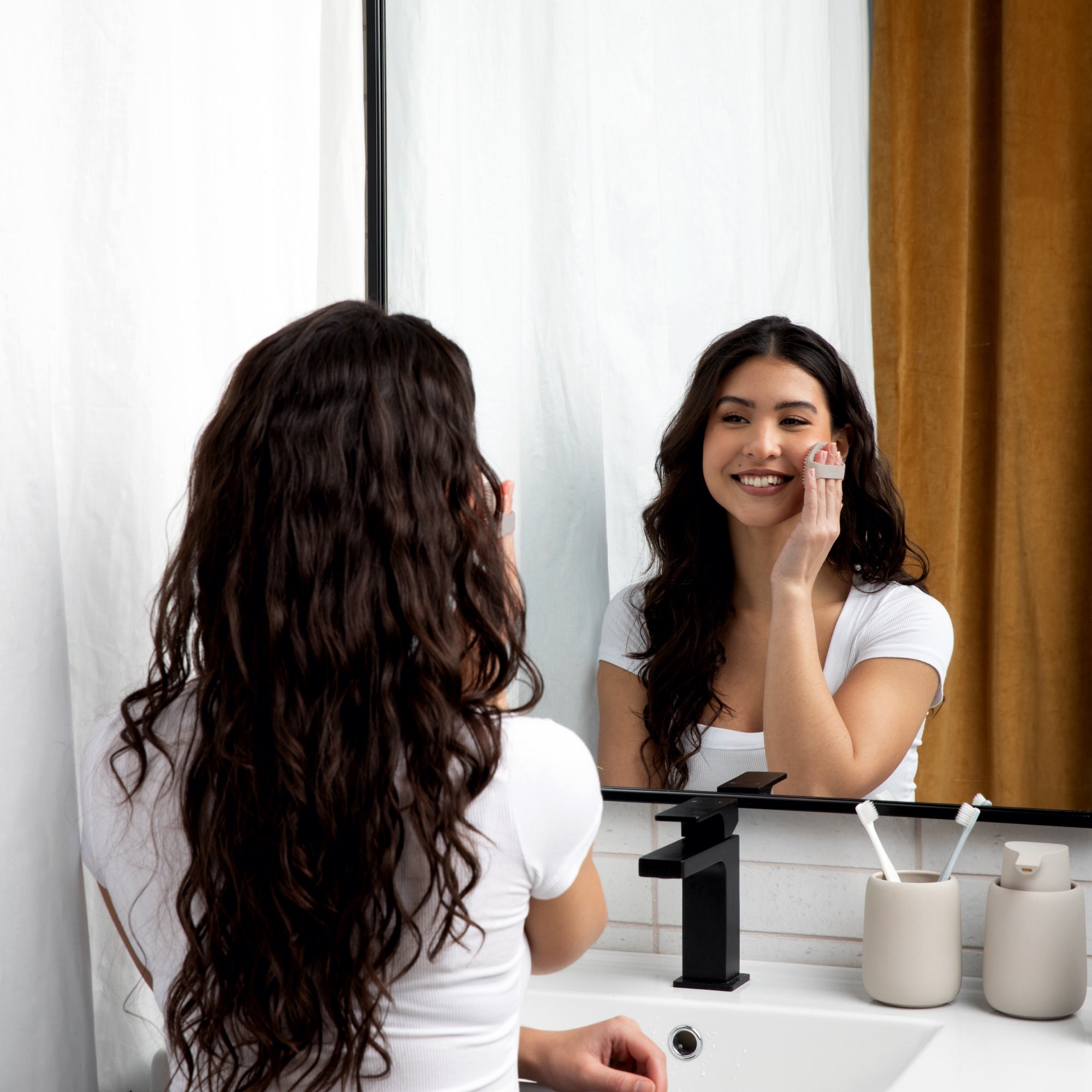 Woman looking at herself in bathroom mirror while using the Sud Scrub face scrubber