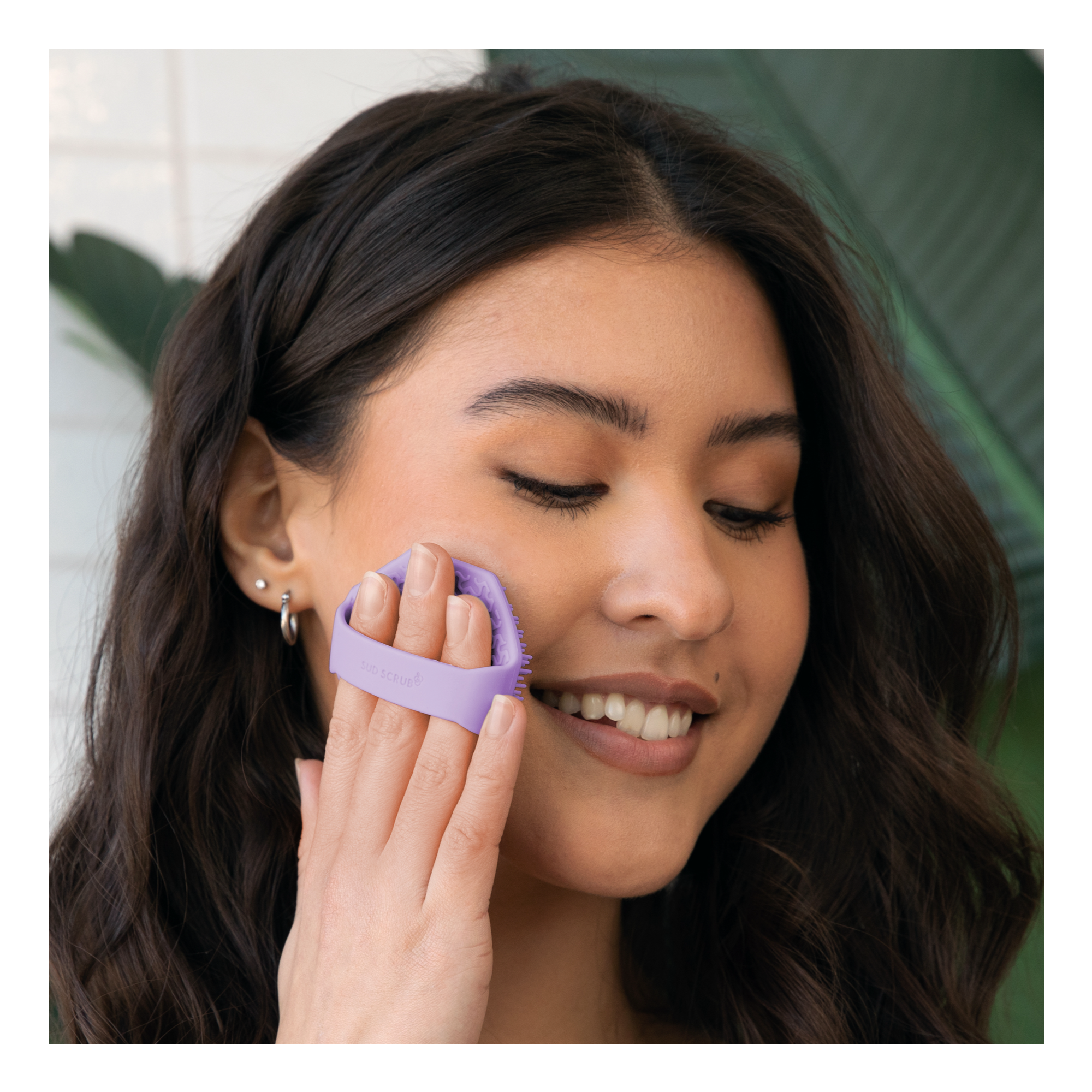 Woman holding purple Sud Scrub Face Scrubber up to her face while smiling.