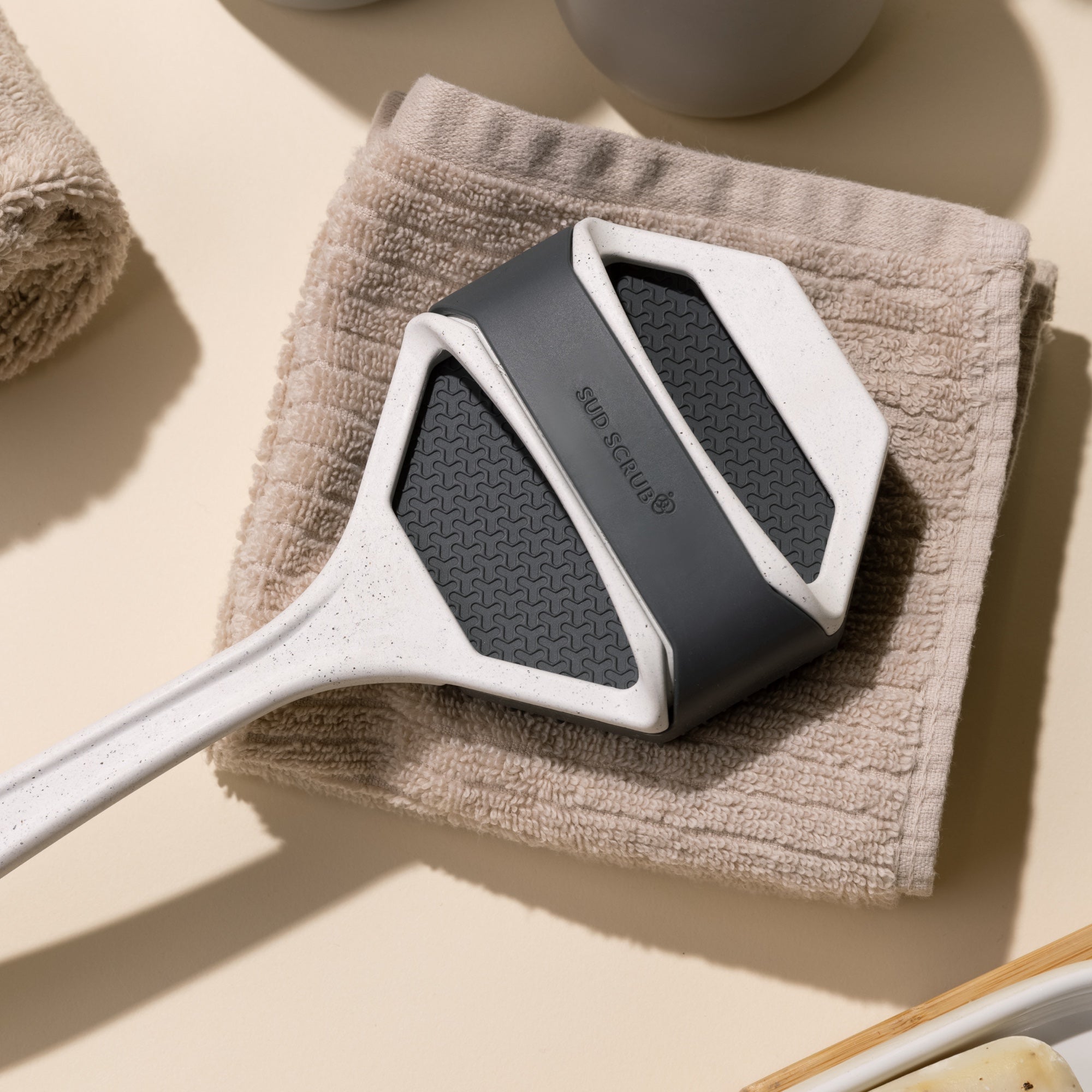 Sud Scrub Body Scrubber fastened onto Sud Scrub Extender Handle in a bathroom setting on a towel