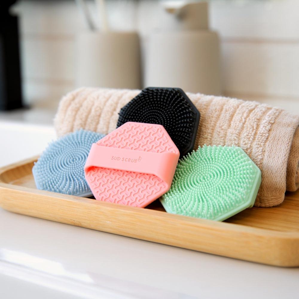 multiple colors of Sud Scrub face scrubbers piled on top of eachother in a bathroom setting